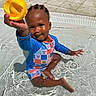 toddler, child, pool, water, swimming, outdoor, sunlight, yellow_scoop, toy, smiling, sitting, wet, playful, blue_clothing, striped_outfit, braids, pavement, tiles, summer, portrait