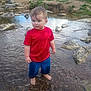 child, toddler, water, creek, rocks, red_shirt, blue_shorts, barefoot, wet_hair, standing, outdoors, nature, stream, shallow_water, pebbles, portrait, candid, summer, curious_face, banks