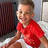 child, boy, smiling, red_shirt, white_shorts, sitting, indoor, blanket, radiator, happy, portrait, cute, hair, face, casual_clothing, furniture, home, cozy, young, kid