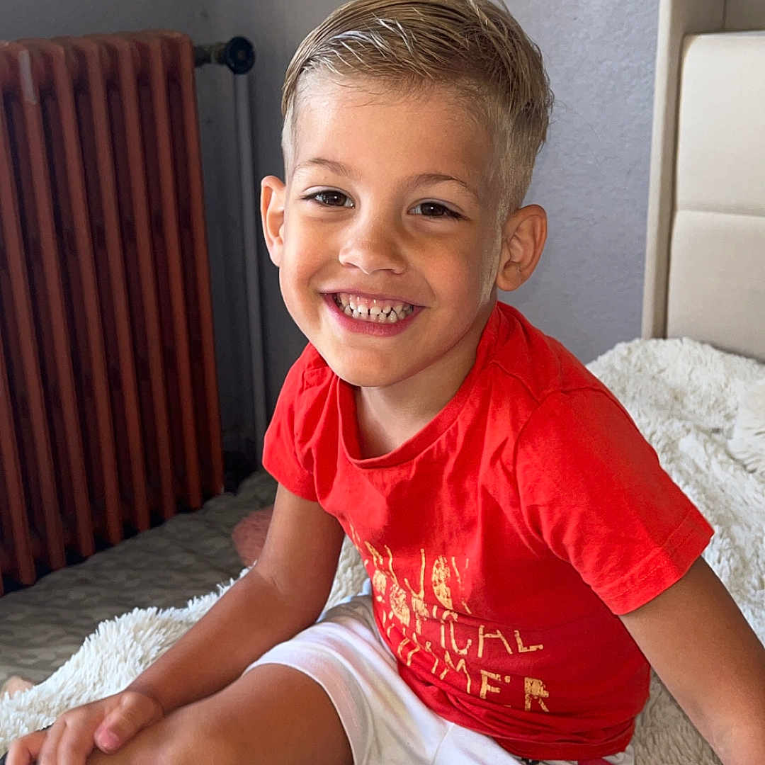 Giulian a rejoint le concours — aidez-le/la à gagner de superbes lots ! child, boy, smiling, red_shirt, white_shorts, sitting, indoor, blanket, radiator, happy, portrait, cute, hair, face, casual_clothing, furniture, home, cozy, young, kid