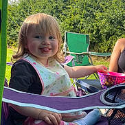 Cassie-Mai is registered to the contest to win money with this photo: toddler, child, outdoor, camping_chair, smiling, tongue_out, bib, bowl, grass, sunlight, greenery, sky, legs, clothing, socks, person, happy, messy_face, nature, playful