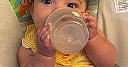Angelica is registered to the contest to win money with this photo: baby, infant, child, portrait, face, eyes, smiling, sitting, cup, drinking, yellow_clothing, striped_clothing, high_chair, baby_seat, animal_pattern, giraffe, monkey, indoor, playful, cute