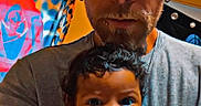 Eziah is registered to the contest to win money with this photo: man, baby, tattoo, cap, facial_hair, earring, indoor, portrait, holding, curly_hair, tshirt, colorful_background, rose_art, expression, person, face, adult, child, skin, hand
