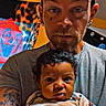 man, baby, tattoo, cap, facial_hair, earring, indoor, portrait, holding, curly_hair, tshirt, colorful_background, rose_art, expression, person, face, adult, child, skin, hand