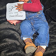 Eziah is registered to the contest to win money with this photo: baby, child, denim_overalls, boots, socks, sign, black_blanket, infant, cute, smiling, portrait, indoor, holding, clothing, person, happy, lying_down, playful, face, expression