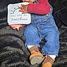 baby, child, denim_overalls, boots, socks, sign, black_blanket, infant, cute, smiling, portrait, indoor, holding, clothing, person, happy, lying_down, playful, face, expression
