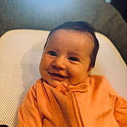 Ella is registered to the contest to win money with this photo: baby, smiling, infant, orange_clothing, cushion, white_background, portrait, happy, child, face, head, closeup, indoors, soft_light, cute, young_child, seated, textile, warm_colors, baby_garment