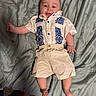 baby, infant, smiling, clothing, shirt, shorts, bed, bedspread, lying_down, happy, cute, child, person, indoors, fabric, skin, footwear, sleepwear, comfortable, playful