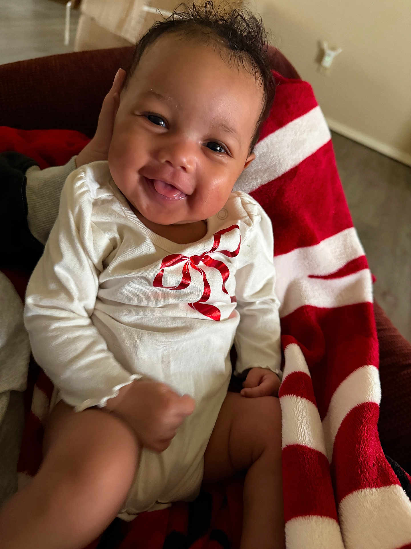 Alani is registered to the contest to win money with this photo: baby, smiling, infant, face, white_clothing, red_bow, blanket, red_and_white, happy, cute, indoors, sitting, person, skin, hand, leg, cozy, portrait, toddler, child
