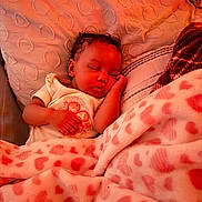 Alani is registered to the contest to win money with this photo: baby, sleeping, blanket, heart_pattern, pillow, cozy, infant, peaceful, soft, bed, child, cute, rest, warm_light, comfort, clothing, portrait, indoor, toddler, relaxation