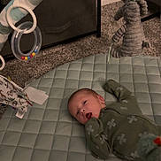 Tucker is registered to the contest to win money with this photo: baby, infant, play_mat, soft_toys, carpet, quilted_mat, toy_ring, green_pajamas, baby_gym, floor, indoor, child, cute, lying_down, plush_toy, baby_clothing, nursery, soft_lighting, home, relaxed