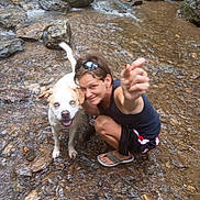 Sabbath is registered to the contest to win money with this photo: person, dog, water, stream, rocks, outdoor, nature, happy, crouching, summer, blue_eyes, shorts, sunglasses, sandals, playful, pet, canine, adventure, smile, shallow_water