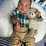 baby, plush_toy, teddy_bear, cushion, plaid_shirt, khaki_pants, brown_shoes, infant, cute, child, toy, bed, striped, bow_tie, bracelet, indoor, person, face, clothing, lying_down