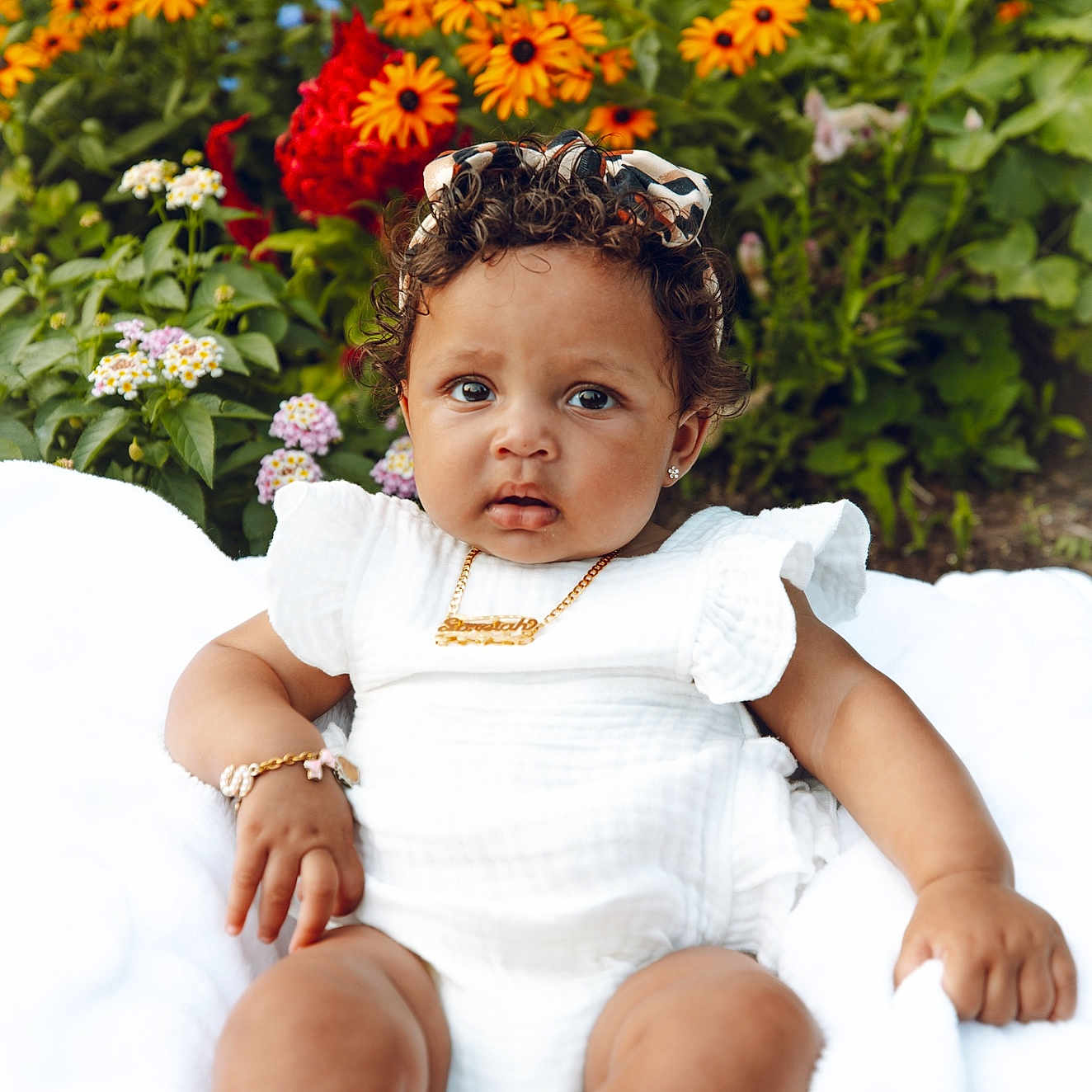 Soreiah joined the competition — help win amazing prizes! baby, child, curly_hair, white_outfit, bracelet, necklace, flowers, orange_flowers, red_flowers, green_leaves, blanket, outdoor, nature, cute, portrait, sitting, infant, garden, headband, expression