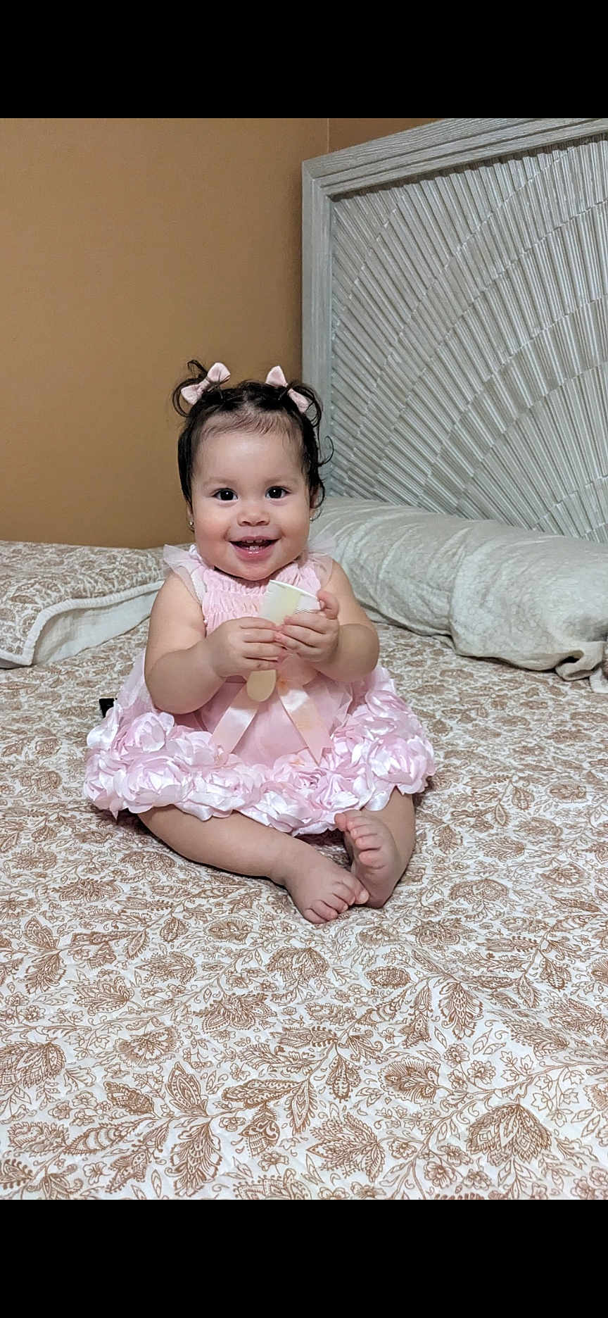 Ana Jazmin is registered to the contest to win money with this photo: baby, child, girl, pink_dress, smiling, sitting, bed, bedspread, patterned, hair_bows, indoor, headboard, happy, cute, infant, portrait, young_child, holding_object, cozy, home