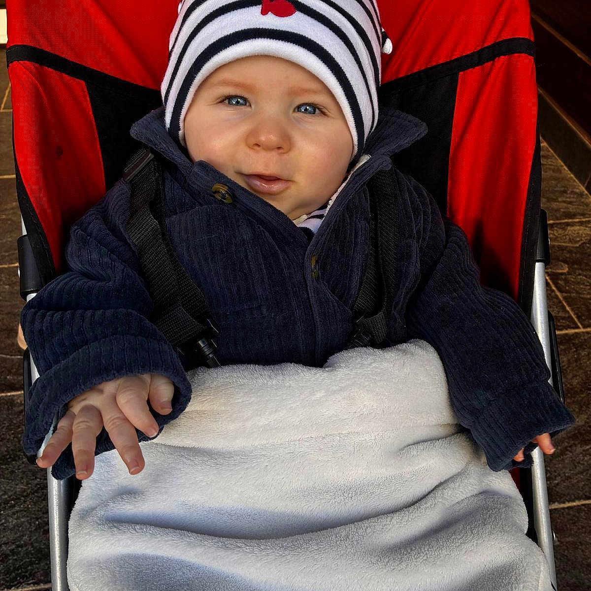 Louis a rejoint le concours — aidez-le/la à gagner de superbes lots ! baby, beanie, blanket, blue_eyes, cheeks, coat, cute, face, fingers, hand, infant, jacket, pavement, portrait, red_backdrop, seat, smile, striped_hat, stroller, winter_clothing