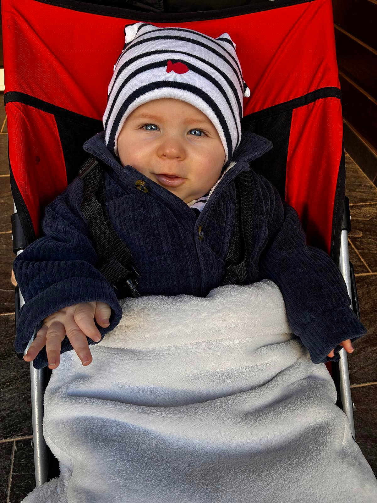Louis a rejoint le concours — aidez-le/la à gagner de superbes lots ! baby, infant, stroller, blanket, striped_hat, beanie, jacket, coat, blue_eyes, smile, hand, fingers, face, cheeks, red_backdrop, seat, pavement, winter_clothing, portrait, cute