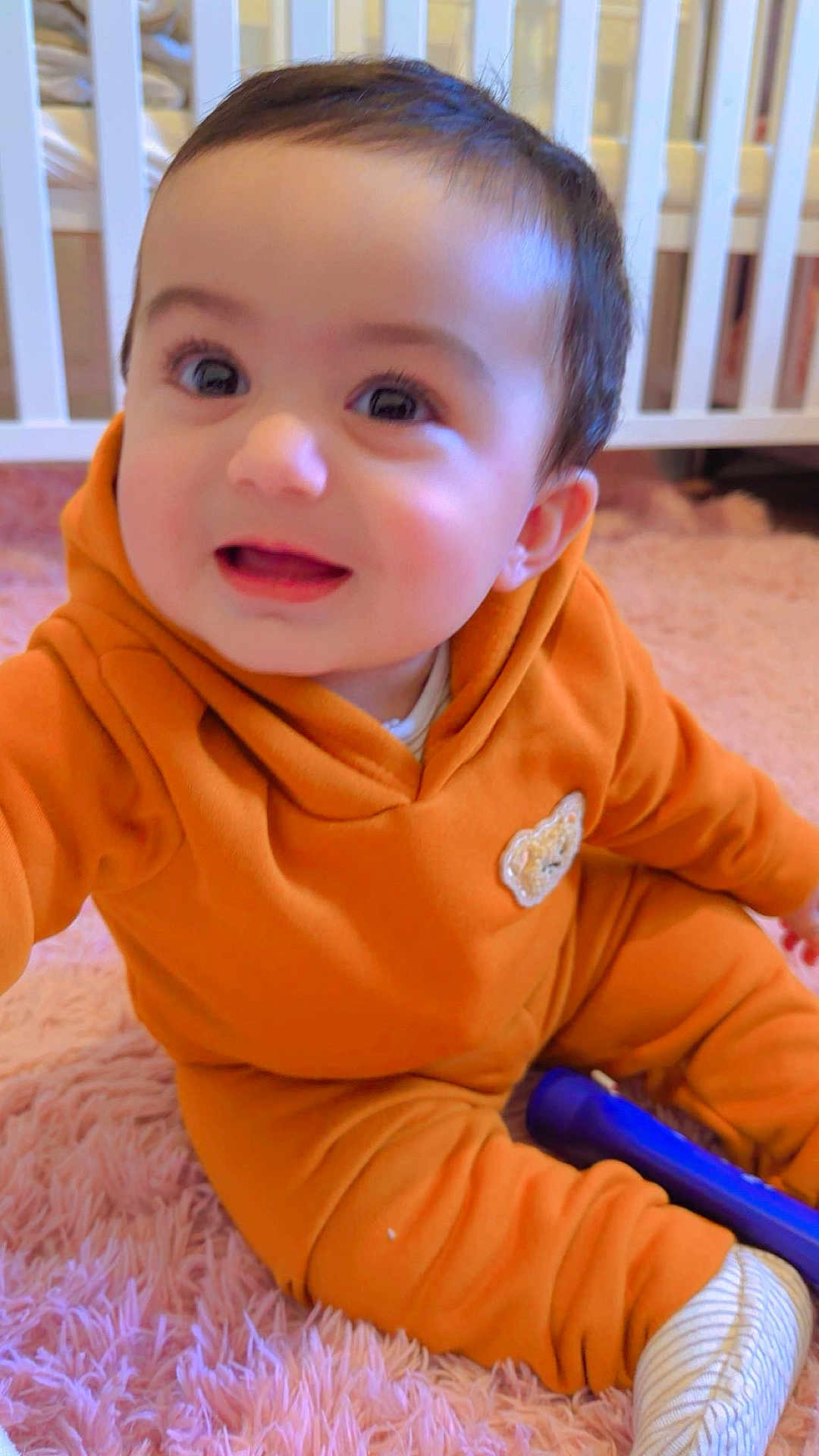 Muhammad is registered to the contest to win money with this photo: baby, child, orange_clothing, sitting, pink_rug, toy, flashlight, crib, indoor, smiling, happy, cute, infant, face, person, cozy, playful, soft, home, portrait