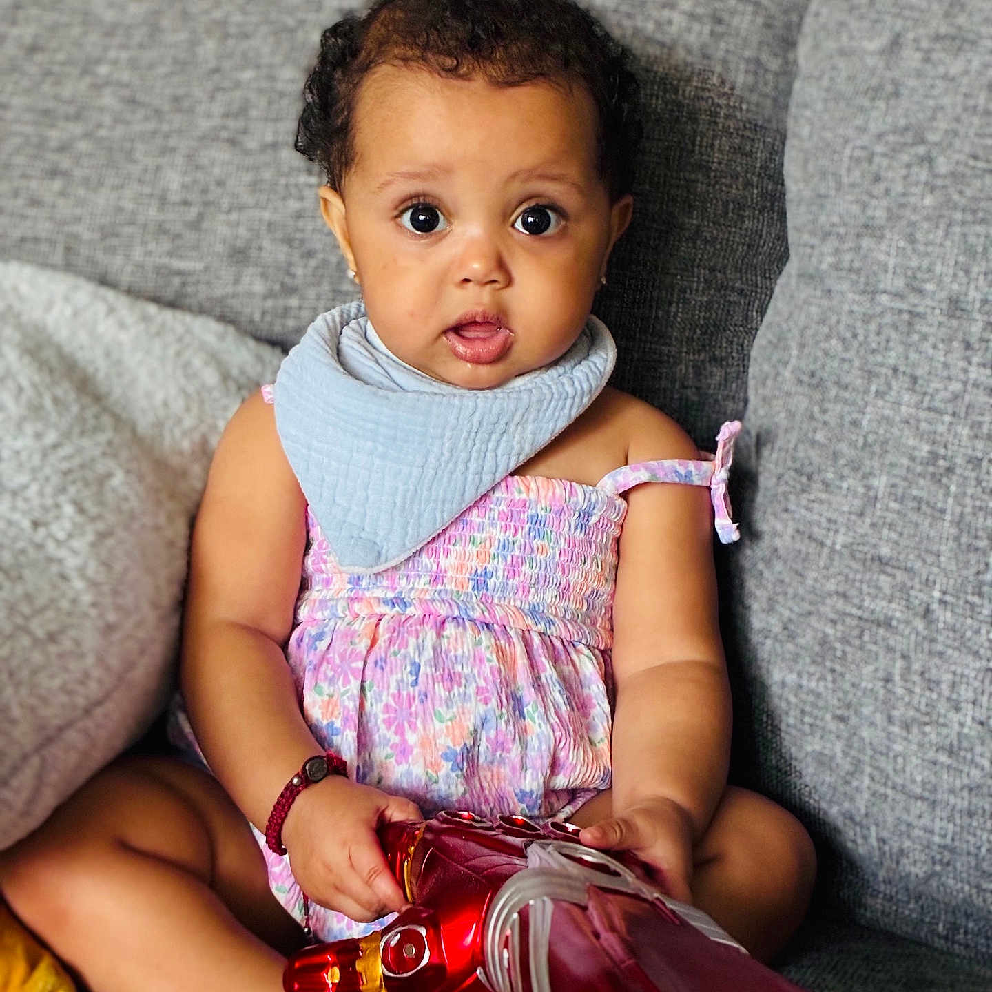 Zoe Zawadi is registered to the contest to win money with this photo: baby, child, infant, sitting, couch, toy, iron_man, dress, bib, curly_hair, barefoot, bracelet, indoors, expression, cute, portrait, person, colorful, playful, home
