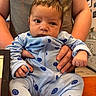baby, infant, onesie, blue_clothing, smiley_faces, adult_hands, tshirt, curly_hair, indoor, table, wooden_table, face, person, holding, furniture, cabinet, decor, expression, cute, child