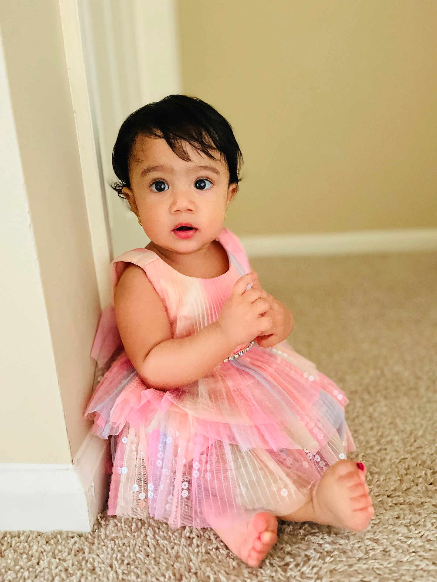 Nitya joined the competition — help win amazing prizes! toddler, child, pink_dress, carpet, indoor, curly_hair, barefoot, sitting, cute, wide_eyes, expression, wall, baseboard, skin, small_earrings, portrait, young_child, innocence, soft_lighting, casual