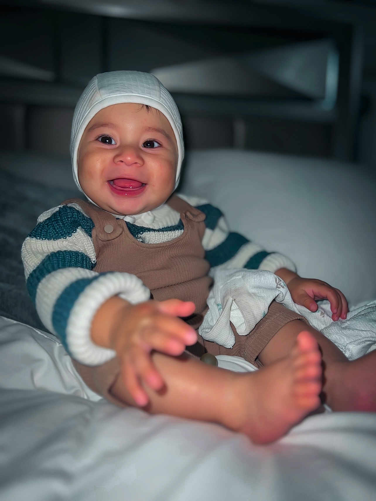 Santiago joined the competition — help win amazing prizes! baby, smiling, child, knit_hat, striped_sweater, overalls, bed, blanket, pillow, happy, indoors, person, cute, skin, foot, hand, portrait, sitting, cozy, warm_clothing
