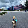child, backpack, hat, road, building, cloudy_sky, asphalt, person, walking, tree, urban, colorful, music_notes, outdoor, daytime, nature, sky, people, adult, casual_clothing