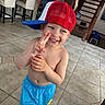 child, toddler, smile, cap, red_cap, blue_cap, barefoot, tile_floor, indoor, home, staircase, bar_stools, shorts, blue_shorts, peace_sign, happy, playful, person, casual, cute
