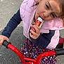 bicycle, bike, candy, casual_clothing, child, daylight, floral_skirt, girl, happy, holding, outdoor, pavement, person, pink_jacket, playful, red, smiling, snack, toy, white_shoes