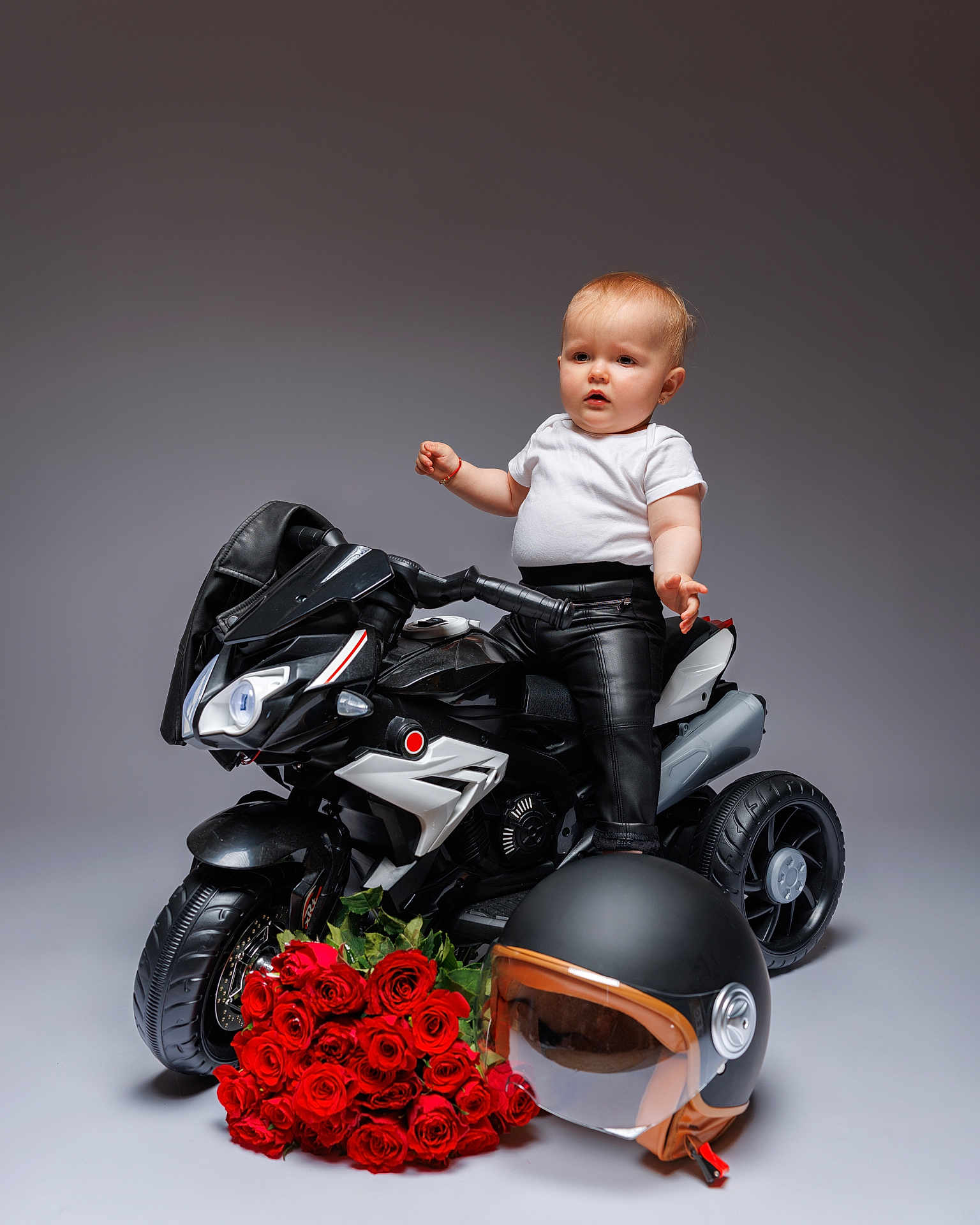 Olivia is registered to the contest to win money with this photo: baby, child, motorcycle, toy, leather_jacket, leather_pants, helmet, roses, bouquet, white_tshirt, black, studio, portrait, pose, floor, wheel, handlebar, expression, indoor, background