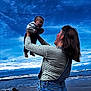 baby, woman, beach, ocean, sky, clouds, smile, joy, holding, denim, striped_shirt, waves, outdoor, portrait, person, happy, child, mother, nature, fun