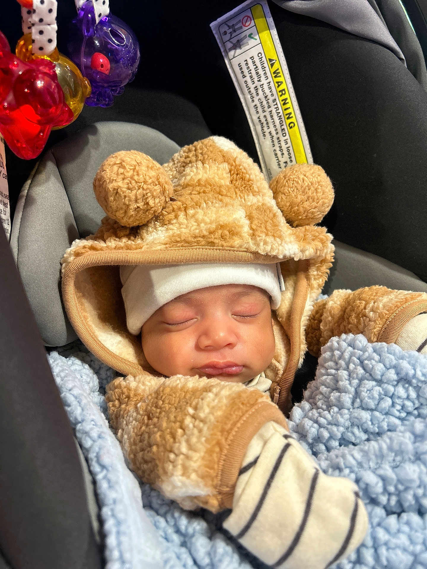 Carlos is registered to the contest to win money with this photo: baby, sleeping, car_seat, blanket, toy, hood, pom_poms, clothing, warm, infant, striped, mittens, soft, cute, child, face, headwear, cozy, indoors, resting