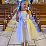 ceremony, child, church, cross_necklace, cute, event, floor_tiles, flower_crown, girl, indoor, lighting, people_in_background, pews, portrait, religious, smile, stone_wall, white_dress, wooden_benches, young