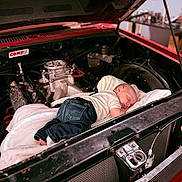 Dominic is registered to the contest to win money with this photo: baby, sleeping, engine_bay, car, vintage_car, blanket, jeans, white_shirt, hand, mechanical_parts, red_car, indoor, workshop, toolbench, cute, infant, resting, soft, metal, automobile