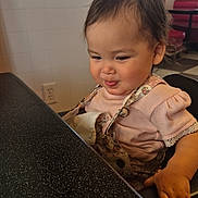 Adeline is registered to the contest to win money with this photo: toddler, child, smirk, pink_clothing, floral_overalls, high_chair, table, speckled_table, hand, face, hair, rosy_cheeks, indoor, restaurant, tile_wall, electrical_outlet, bench_seating, expression, close_up, portrait