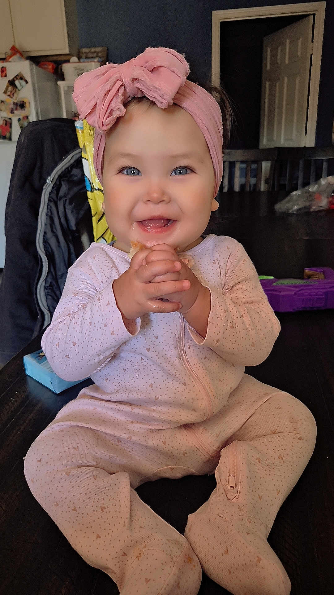 Adeline joined the competition — help win amazing prizes! baby, infant, child, smiling, headwrap, bow, onesie, sitting, hands, food, drooling, blue_eyes, rosy_cheeks, teeth, table, indoor, kitchen, refrigerator, toy, portrait