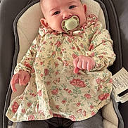 Myla a rejoint le concours — aidez-le/la à gagner de superbes lots ! baby, baby_chair, child, clothing, comfort, cushion, cute, expression, face, floral_dress, hands, indoor, infant, pacifier, pants, person, portrait, seated, soft_lighting, young_child