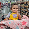 Aria is registered to the contest to win money with this photo: child, toddler, baby, smiling, shopping_cart, grocery_store, supermarket, cashews, snack, blanket, yellow_overalls, purple_shirt, curly_hair, earrings, portrait, indoor, shopping, sitting, happy, food_display