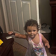 Aria is registered to the contest to win money with this photo: toddler, child, baby, standing, smiling, portrait, indoor, home, pink_dress, cartoon, clothing, curly_hair, face, eyes, arms, feet, carpet, door, furniture, table
