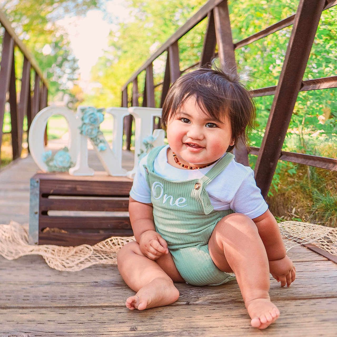 Matthyus is registered to the contest to win money with this photo: baby, bridge, child, cute, daylight, face, greenery, happy, infant, nature, one, outdoor, person, portrait, romper, sitting, smiling, sunlight, wood, wooden_floor