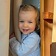 Adèle participe au concours pour gagner de l'argent avec cette photo : baby, blonde_hair, blue_eyes, child, closeup, clothing, curtain, cute, face, hair_bow, happy, indoor, long_sleeve, natural_light, person, playful, portrait, smiling, toddler, wood