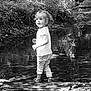 child, toddler, water, creek, nature, outdoor, reflection, plants, trees, rocks, shoes, pants, shirt, black_and_white, curly_hair, smile, standing, daylight, forest, playful