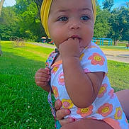 Cimani is registered to the contest to win money with this photo: baby, child, outdoor, grass, headband, yellow_bow, onesie, flower_pattern, hand_in_mouth, greenery, sunny_day, park, trees, blue_sky, cute, infant, person, nature, summer, playground