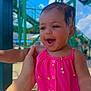 baby, child, pink_clothing, smiling, outdoor, daylight, water_park, green_structure, hand, happy, playful, summer, sunlight, infant, portrait, clothing, person, face, background, fun