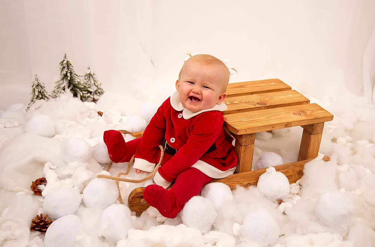 Darcie-Rose joined the competition — help win amazing prizes! baby, child, red_clothing, wooden_sled, snowballs, pinecones, winter, holiday, festive, smiling, indoor, decorations, white_background, cute, toddler, snow, playful, joyful, portrait, seasonal