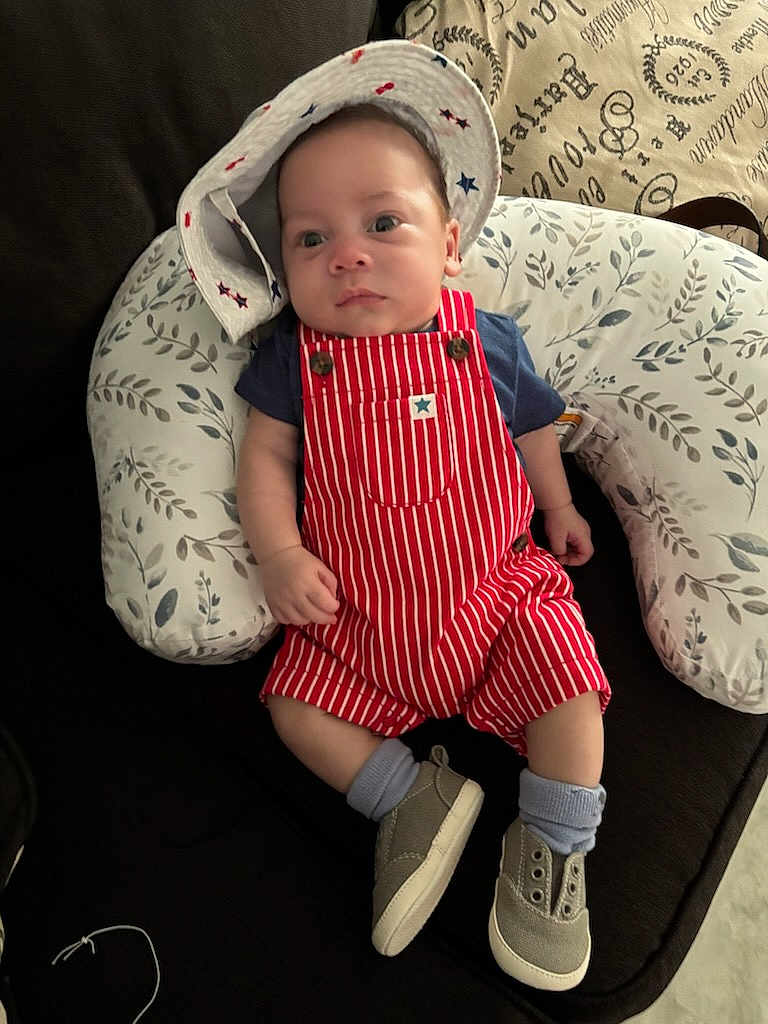 Kaysen joined the competition — help win amazing prizes! baby, infant, hat, romper, striped_clothing, shoes, socks, pillow, couch, patterned, face, person, clothing, indoor, cute, child, seated, footwear, relaxed, young