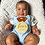 baby, infant, smile, superman_logo, blue_overalls, wooden_sign, four_months, blanket, soft_texture, cute, happy, portrait, child, clothing, sitting, person, young_child, indoors, closeup, baby_girl