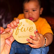 Dylan is registered to the contest to win money with this photo: baby, child, hand, wooden_plaque, yellow_shirt, bracelet, person, infant, five_months, holding, close_up, indoor, focus, skin, fingers, cute, expression, portrait, sitting, celebration
