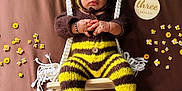 Dylan is registered to the contest to win money with this photo: baby, bee, black, brown_background, child, costume, cute, decorations, feet, flowers, hands, infant, knitwear, plaque, portrait, striped, swing, three_months, wooden, yellow
