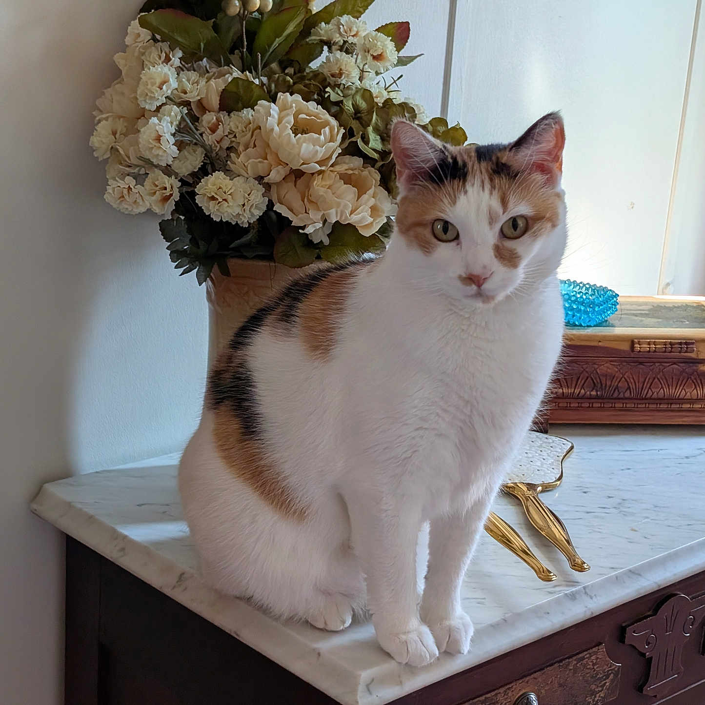 Peppermint Patty joined the competition — help win amazing prizes! cat, calico_cat, flower_bouquet, marble_surface, wooden_cabinet, decorative_object, hand_mirror, indoor, pet, feline, curious, sitting, closeup, vintage, ornate, tabletop, white_wall, green_leaves, soft_light, domestic